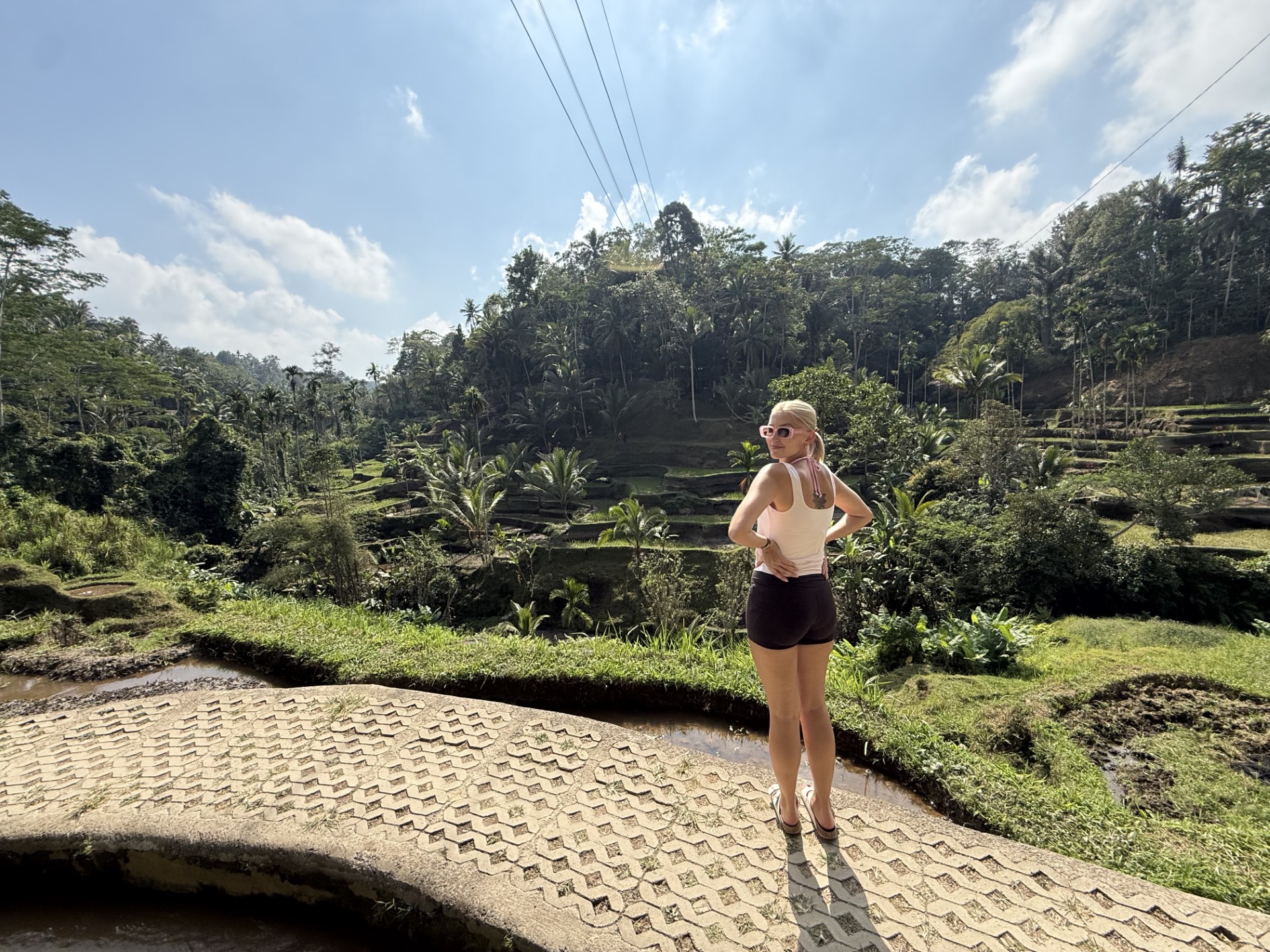 A blond woman stands on a bridge in front of green flora