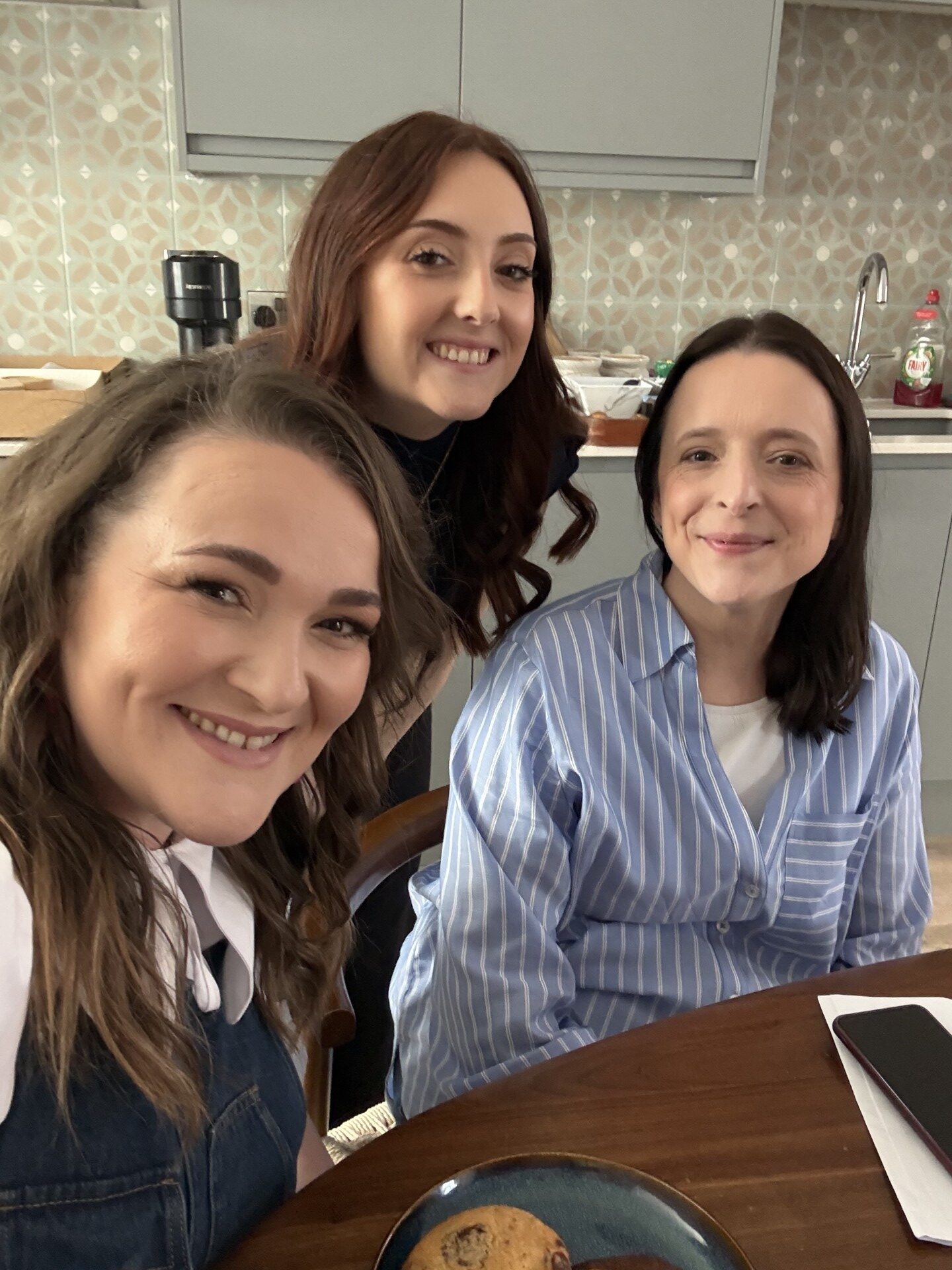 Three brunette women smiling at the camera, sitting in front of a kitchen table