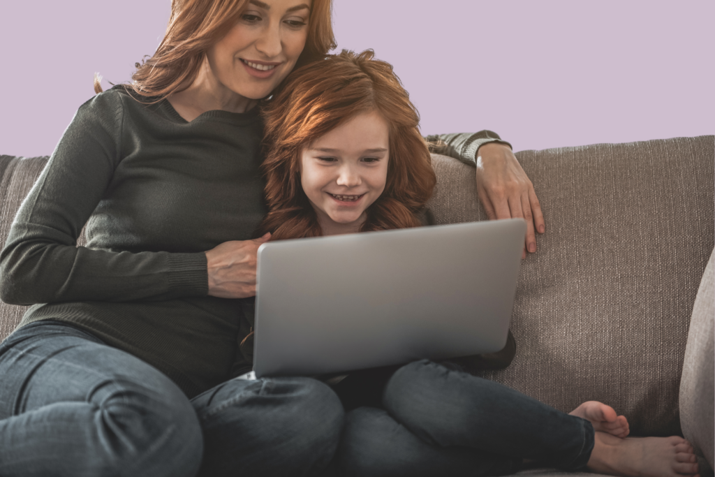 A mother and daughter smile as they look at a laptop while sitting on the sofa together.