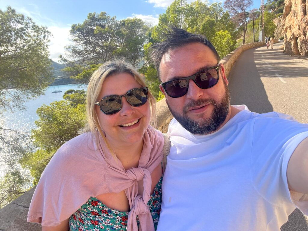 A man and a woman pose for a selfie in the sun in front of greenery and water