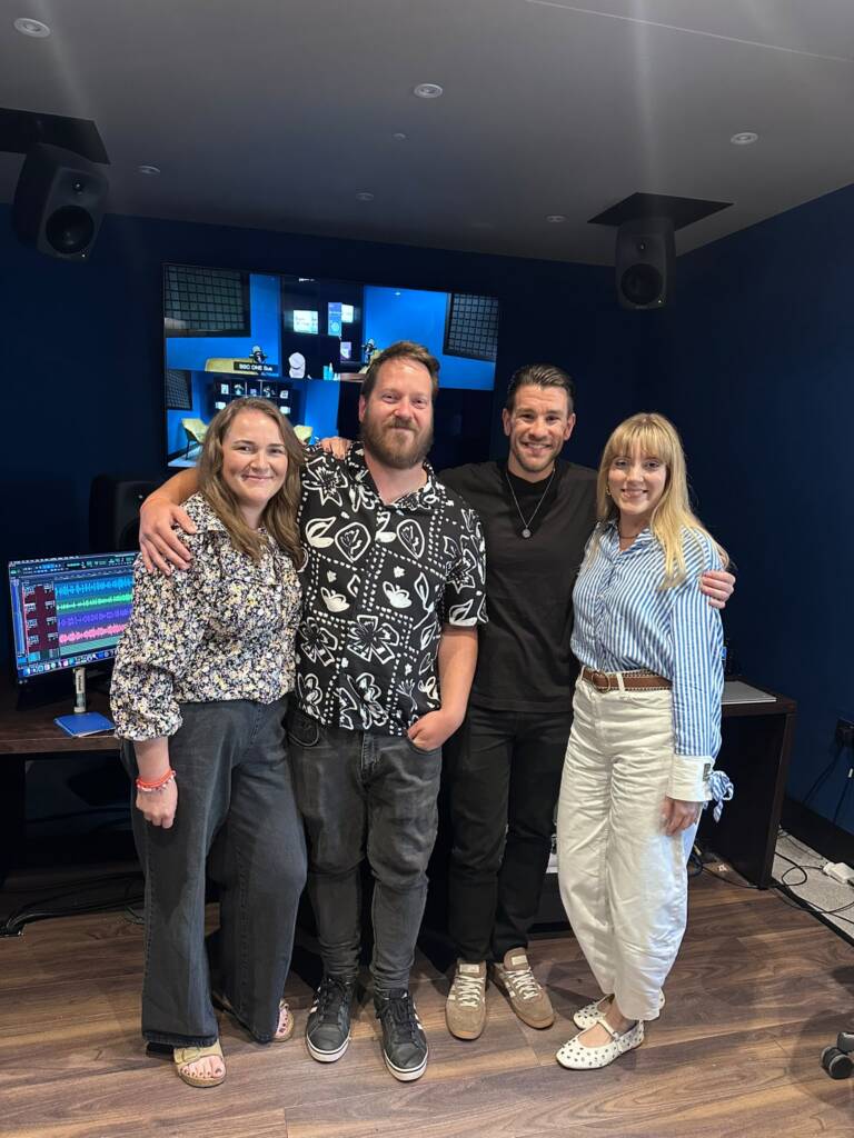Two couples smile with their arms around each other, in front a computer inside a recording studio. 