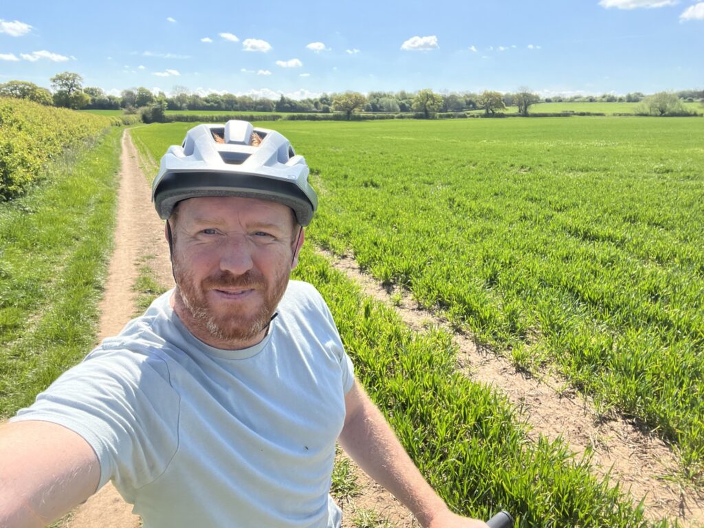 A red-haired man with a bike helmet on stands in a green field
