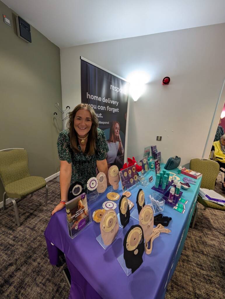 A brunette woman in a green dress smiles as she leans over in front of a table covered in ostomy products 