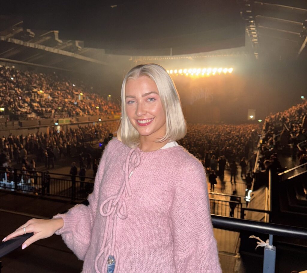 A woman in a pink jumper stands in front of a large crowd at Wembley Arena