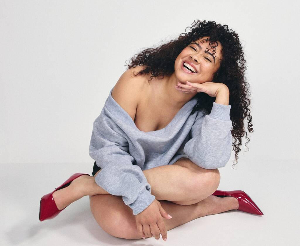 A woman with curly hair sits cross legged on the floor smiling, in a grey jumper and reed shoes.