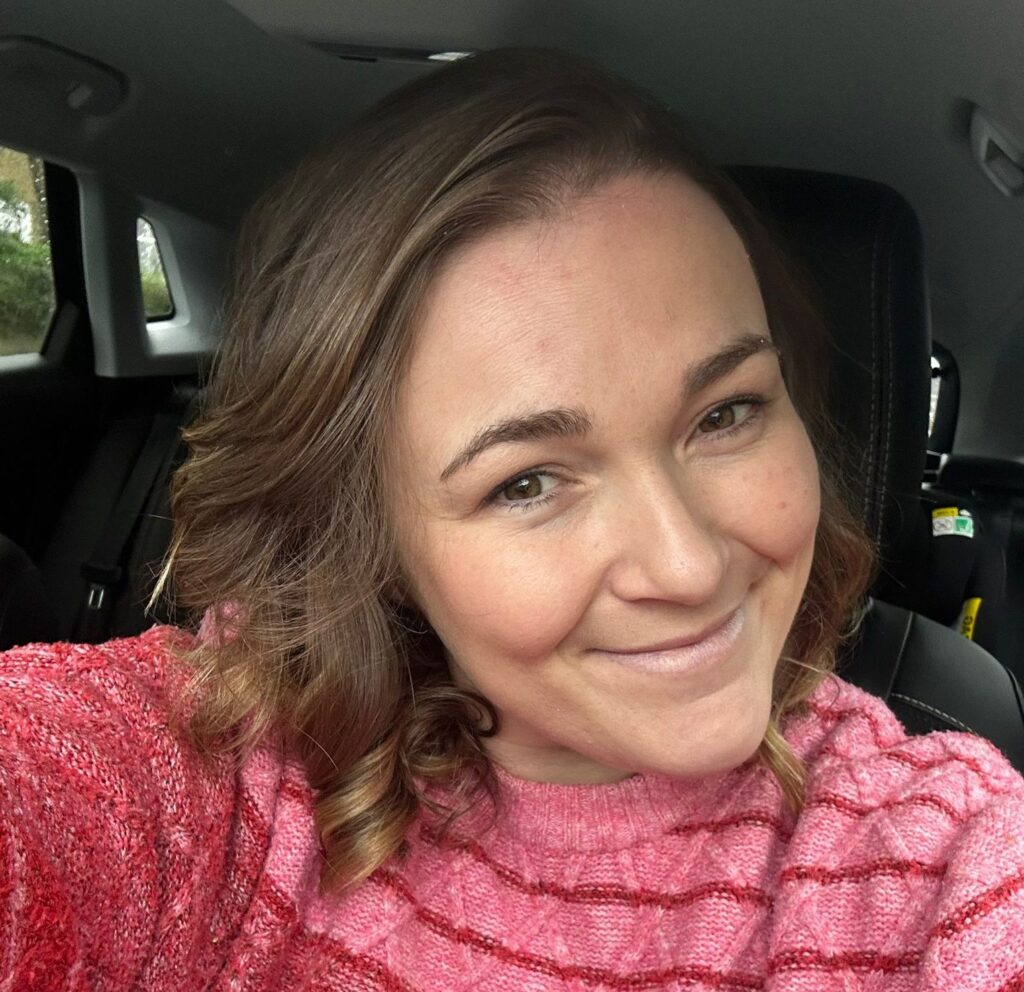 A woman in a pink jumper smiles at the camera while sitting in her car.