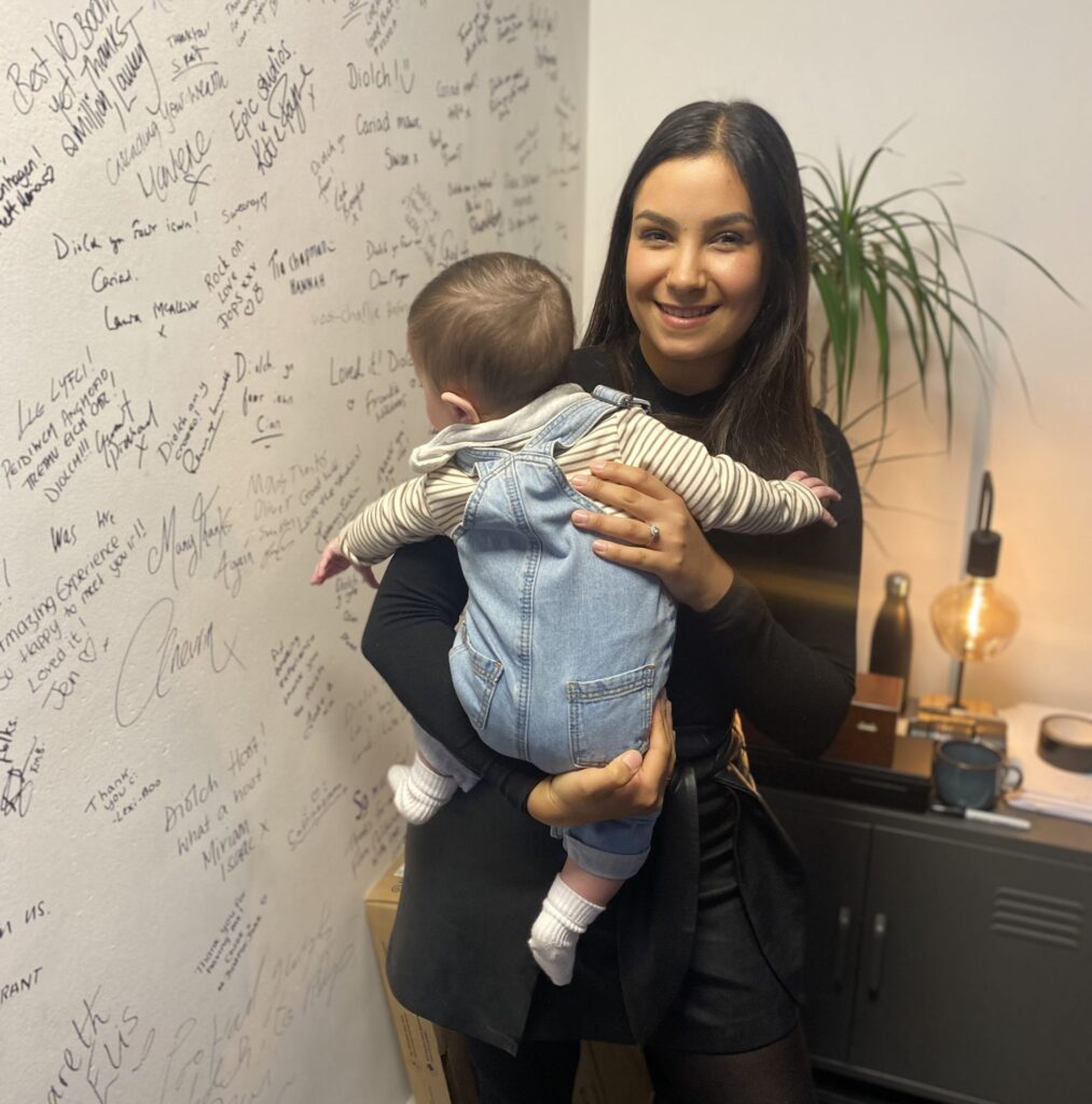 A dark-haired woman holds a baby, stood next to a wall covered in writing