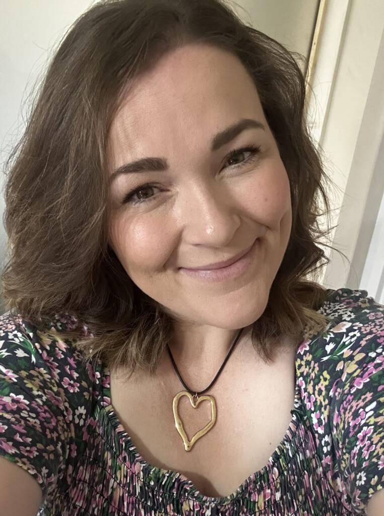 A brown-haired woman smiles at the camera wearing a pink and green floral dress