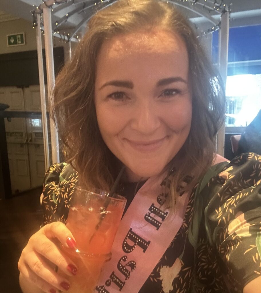 A woman smiles at the camera wearing a pink sash and holding a drink