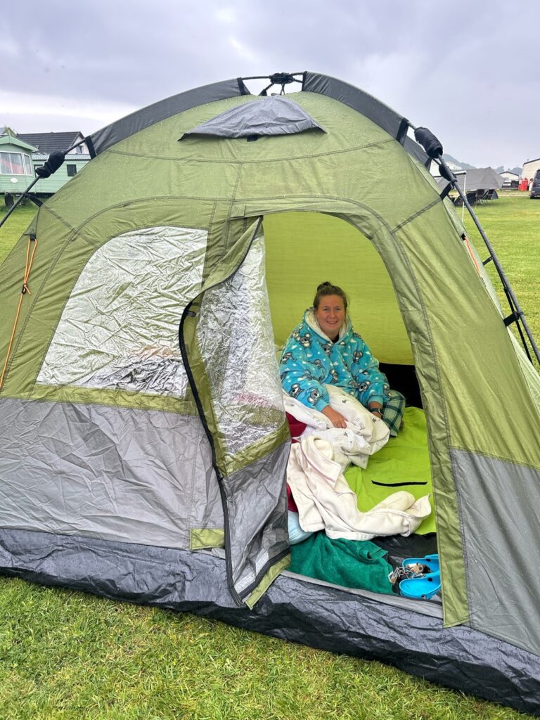 A woman smiles at the camera, wearing a dryrobe and sitting inside a tent in the middle of the field.
