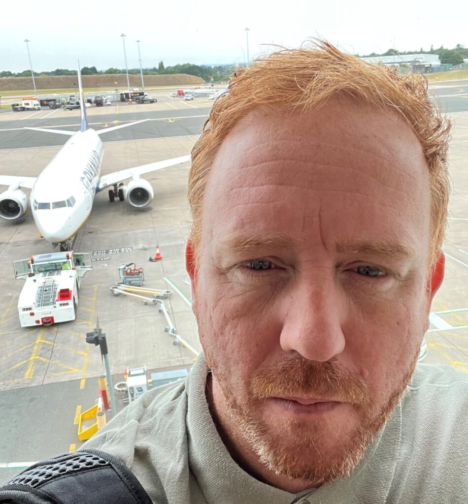 A red-haired man takes a selfie in front of a passenger jet
