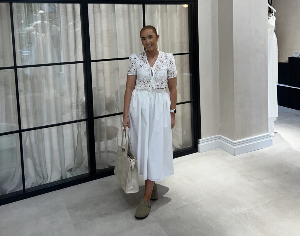 A woman poses in front of glass door, wearing a white wedding dress and holding a white bag