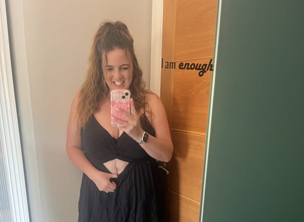A red-haired woman poses in front of a door with her stoma bag showing, with the text "I am enough" on screen