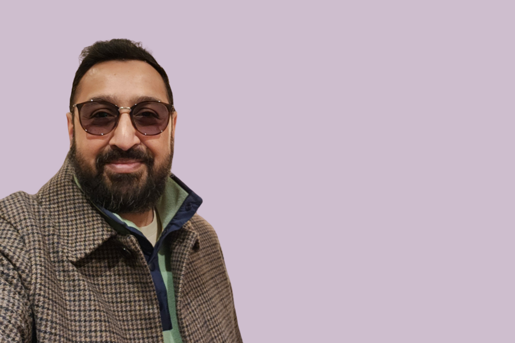 A man with a beard and sunglasses smiles in front of a purple background