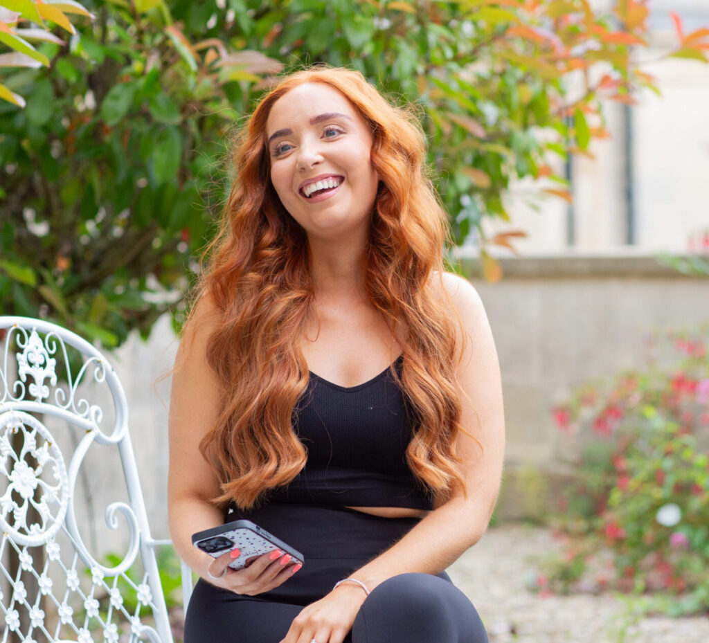 A red-headed woman smiles as she holds her phone, sitting on a garden chair in front of a tree