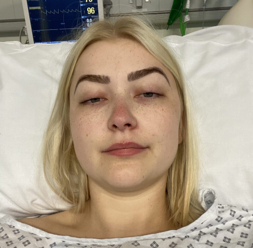 A selfie of a blonde woman lying in bed, wearing a hospital gown