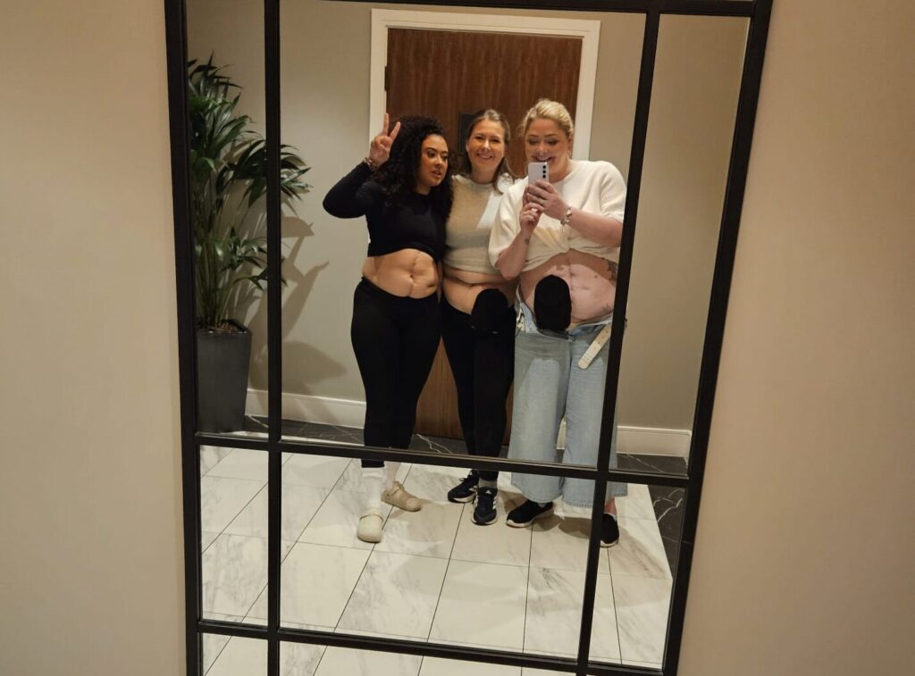 Three women with ostomy bags pose in a full-mirror selfie.