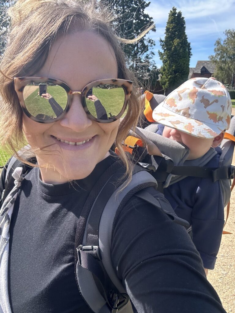 A woman in sunglasses smiles as she walks through the countryside with a baby on her back