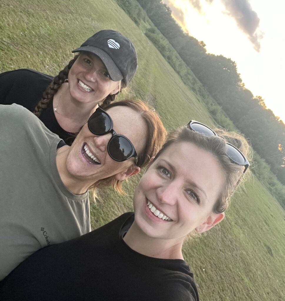 Three women smile looking at the camera, standing in a field