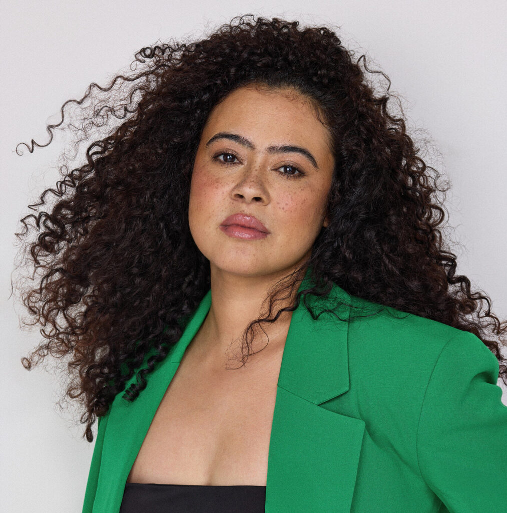A woman with dark, curly hair and a green jacket poses for the camera.