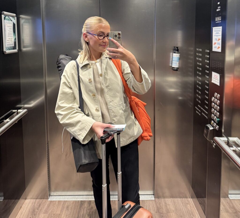 A blonde woman takes a selfie in a lift holding a suitcase and a bag.