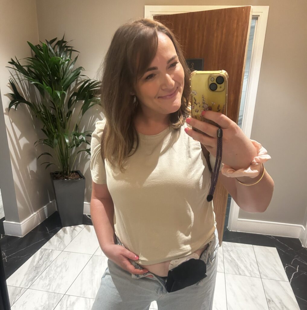 A woman stands in the mirror with her jeans unzipped, showing her ostomy bag