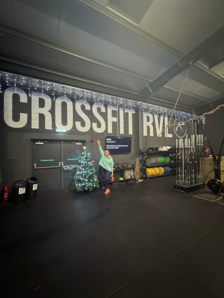 A young woman in a green jumper open her arms and poses underneath a sign saying "CROSSFIT RVL"
