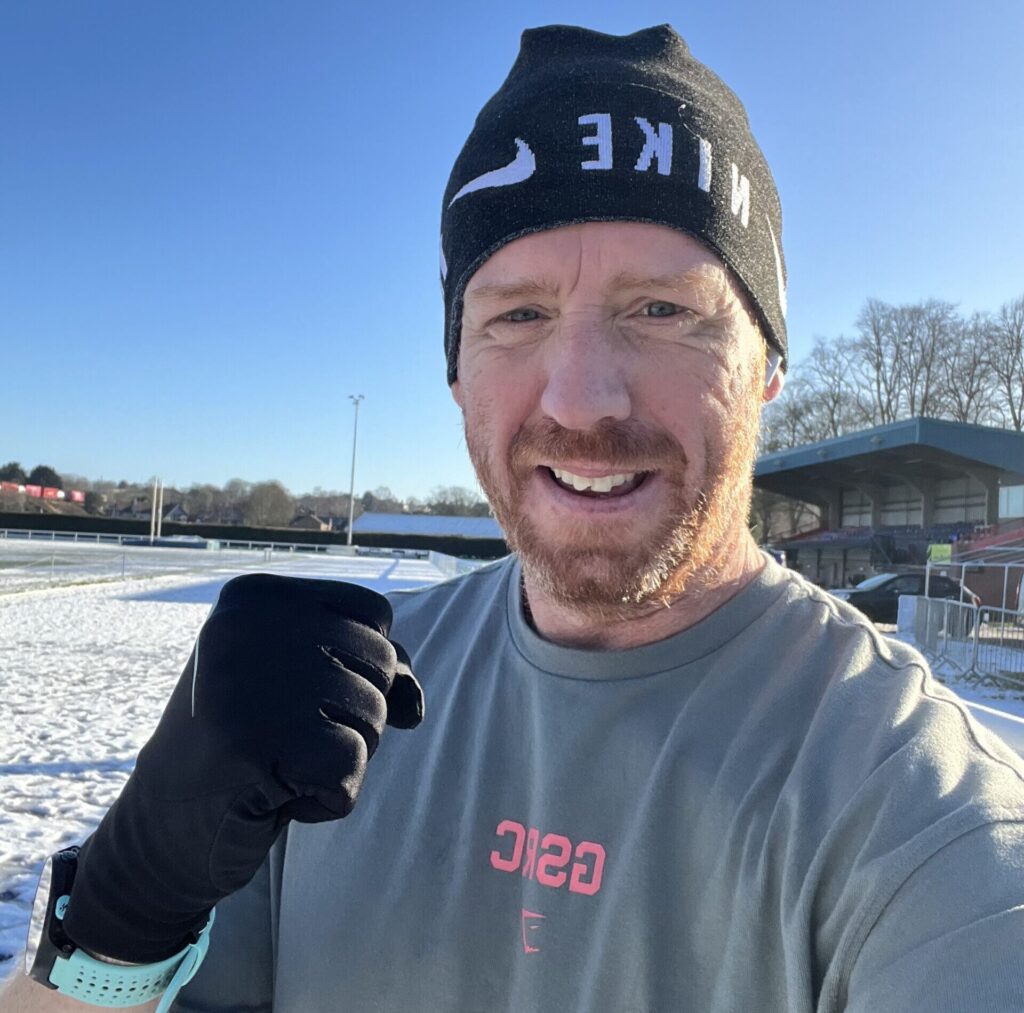 A red-haired man in a winger hat and gloves poses with his first up in the snow.