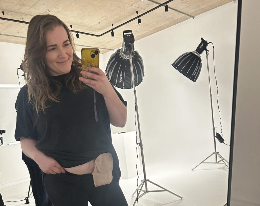 A brunette woman poses with her stoma bag out, taking a selfie backstage at a photoshoot