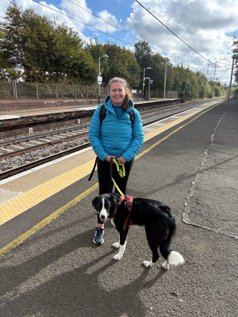 A woman in a blue coat holds a dog on a lead, as they wait at a train station.
