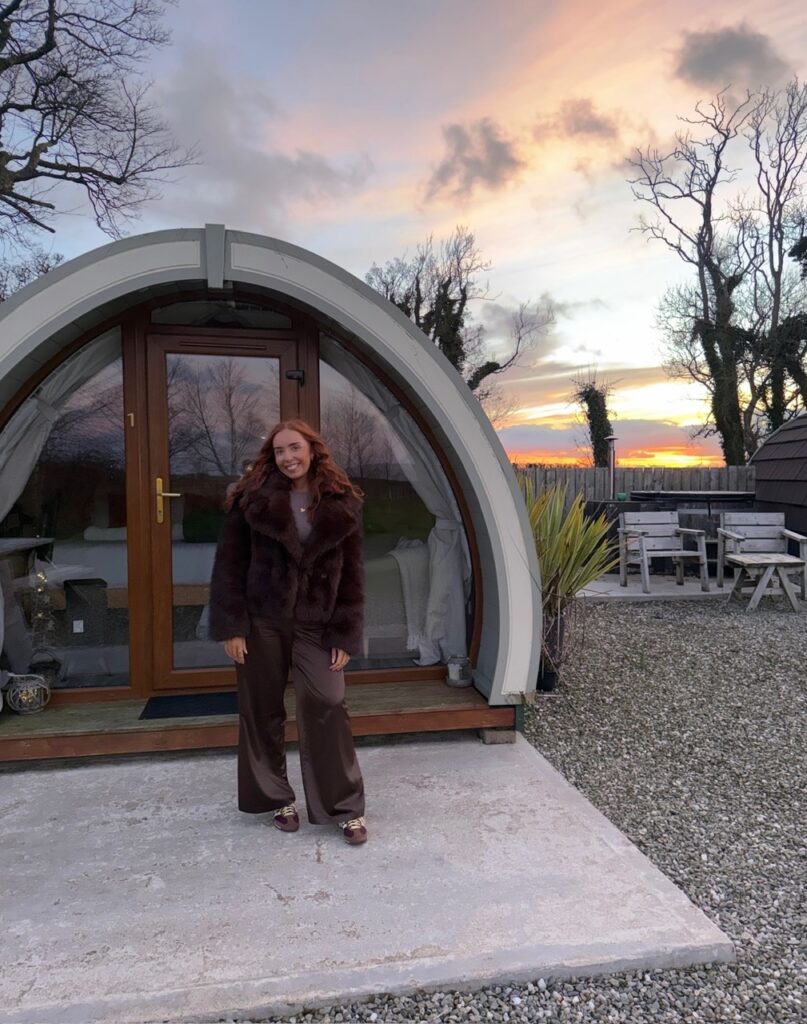 A red-haired woman stands in front of a wood lodge at sunset