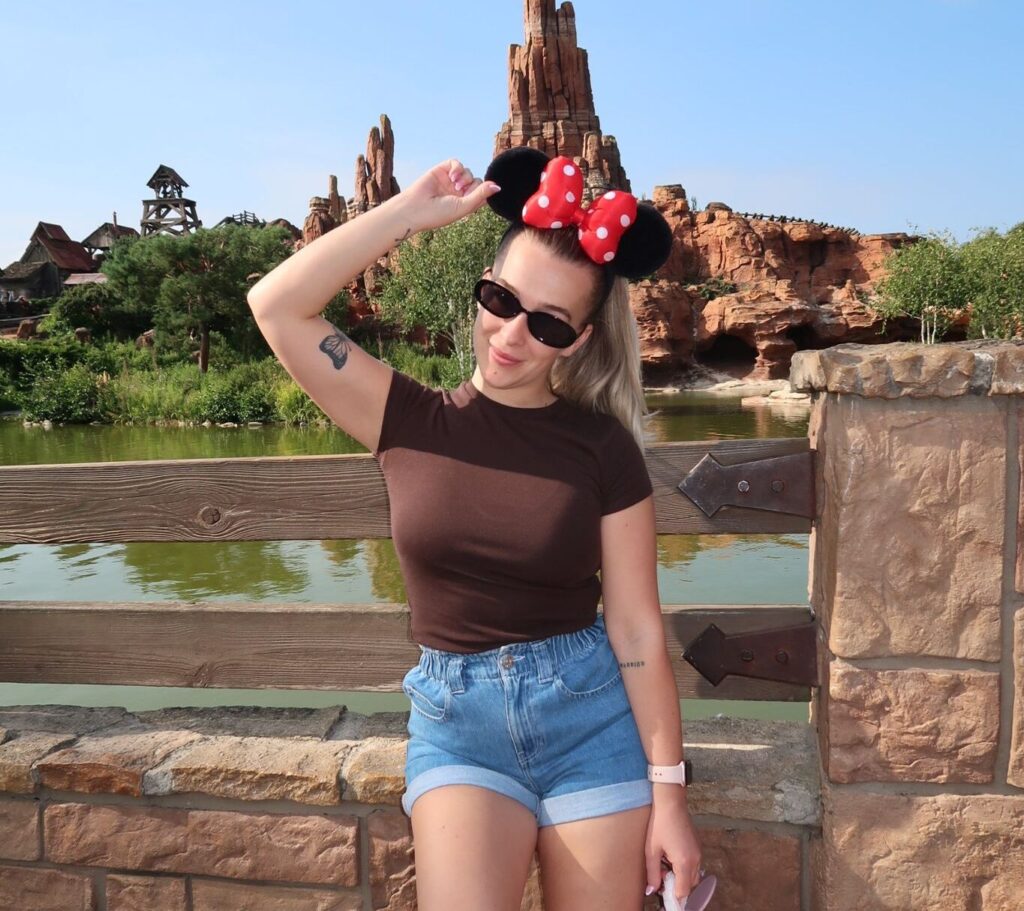 A man in brown shirt and jean shorts poses in Disneyland wearing Minnie Mouse ears