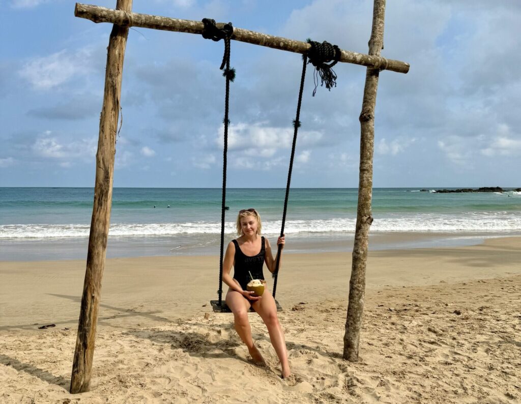 A woman sits on a swing on a beach in Sri Lanka, wearing a black swimsuit and holding a coconut