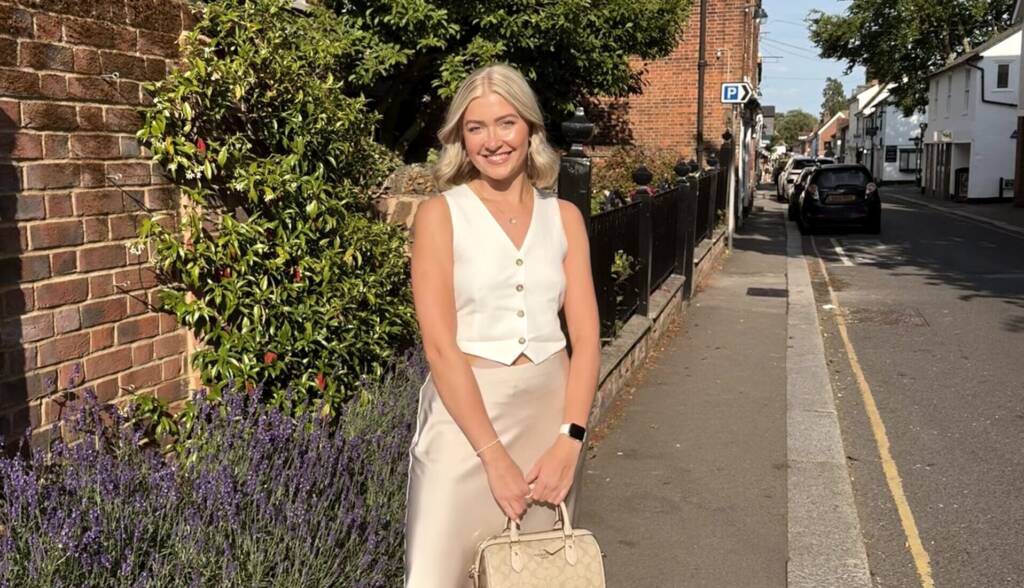 A blonde woman in a waistcoat and a skirt smiles while she holds a bag, standing at the end of a street in the sunshine
