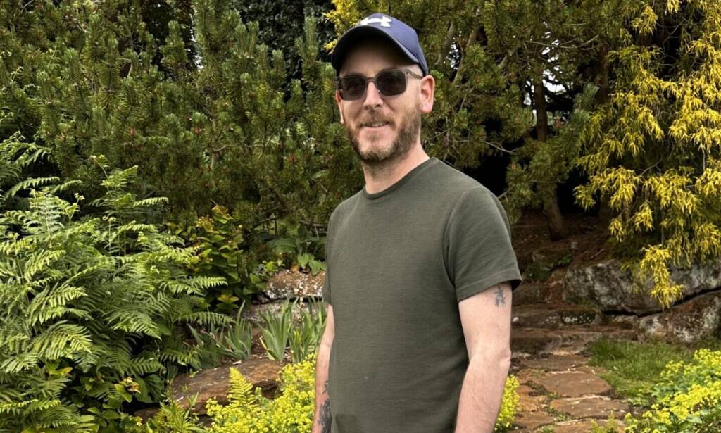 A man in a hat and glasses stands among green shrubbery