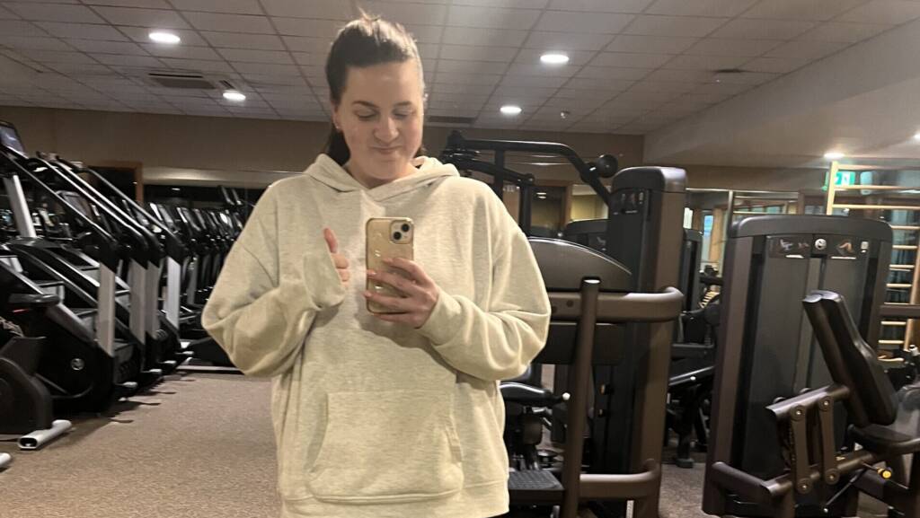 A woman in a white hoodie and black leggings smiles as she takes a selfie in the gym