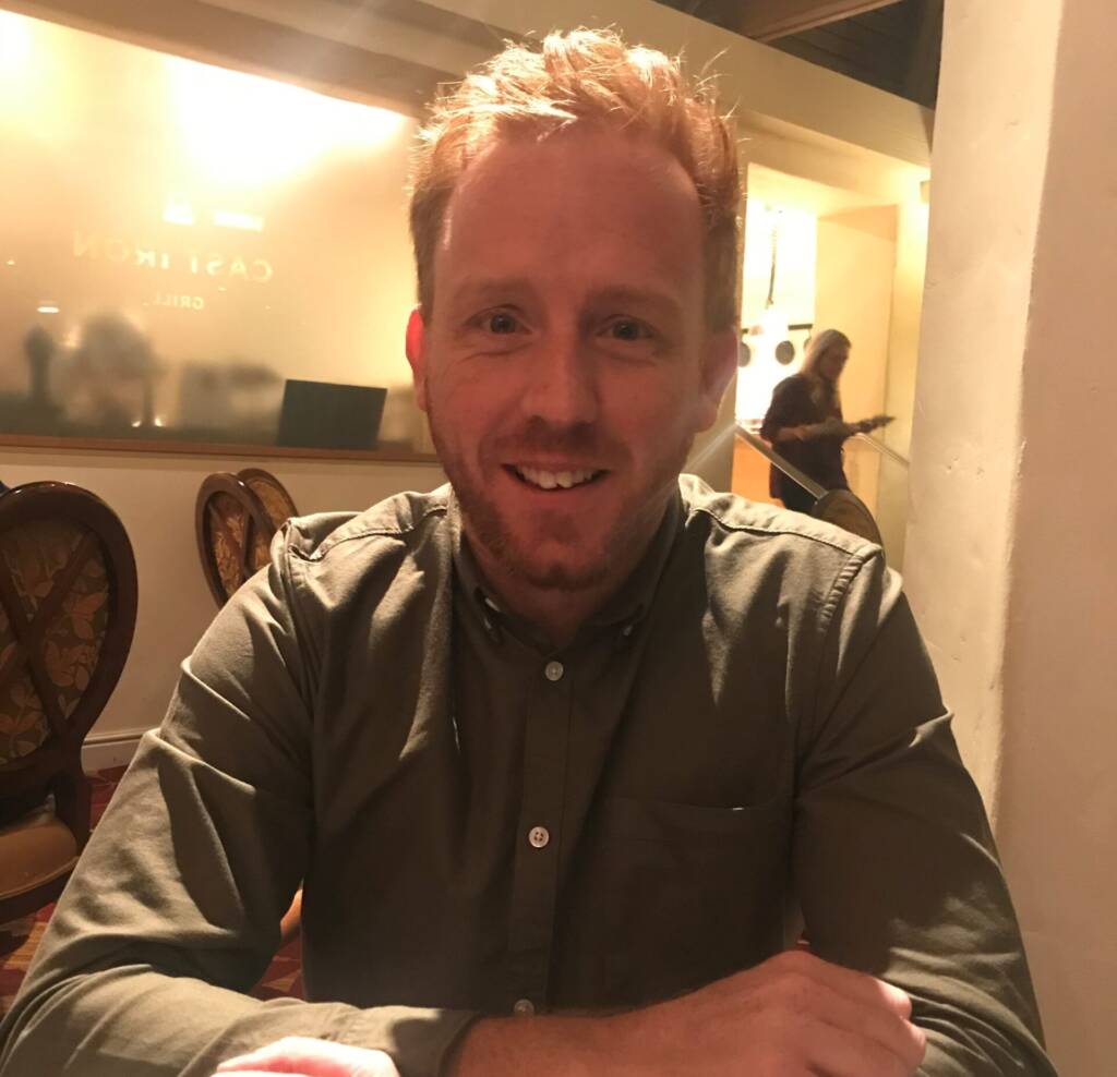 A red-haired man smiles with his hands crossed, sitting in a low-light restaurant