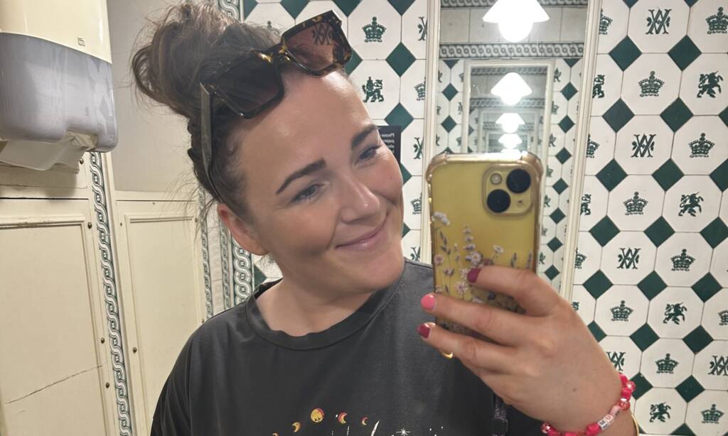 A woman in a grey t-shirt with a sun on it poses in a bathroom mirror, taking a selfie while exposing her ostomy bag.
