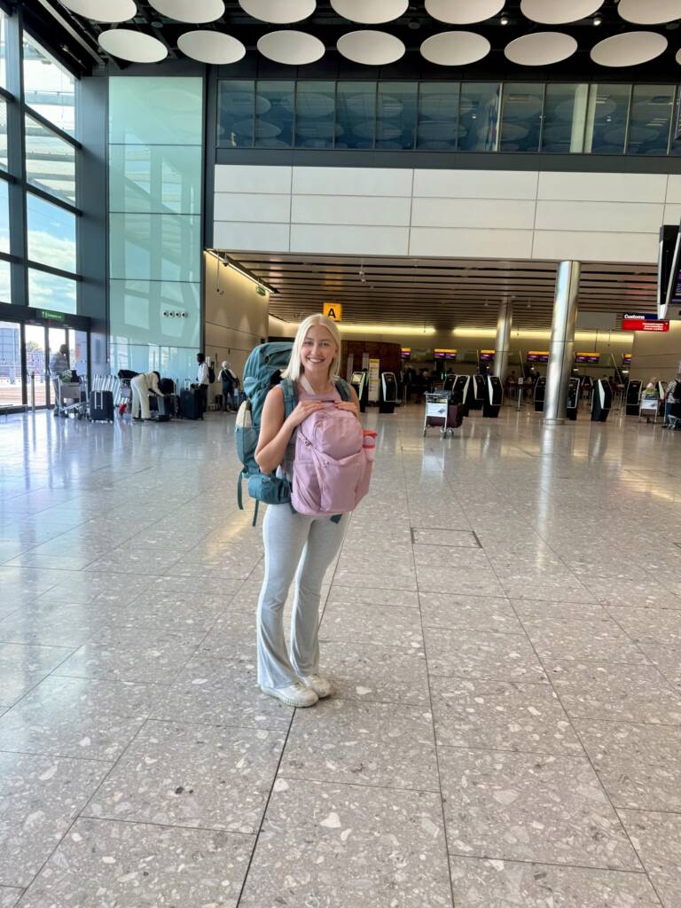 A blonde woman stands in an airport, with a blue backpack on her back and a pink bag on her front.