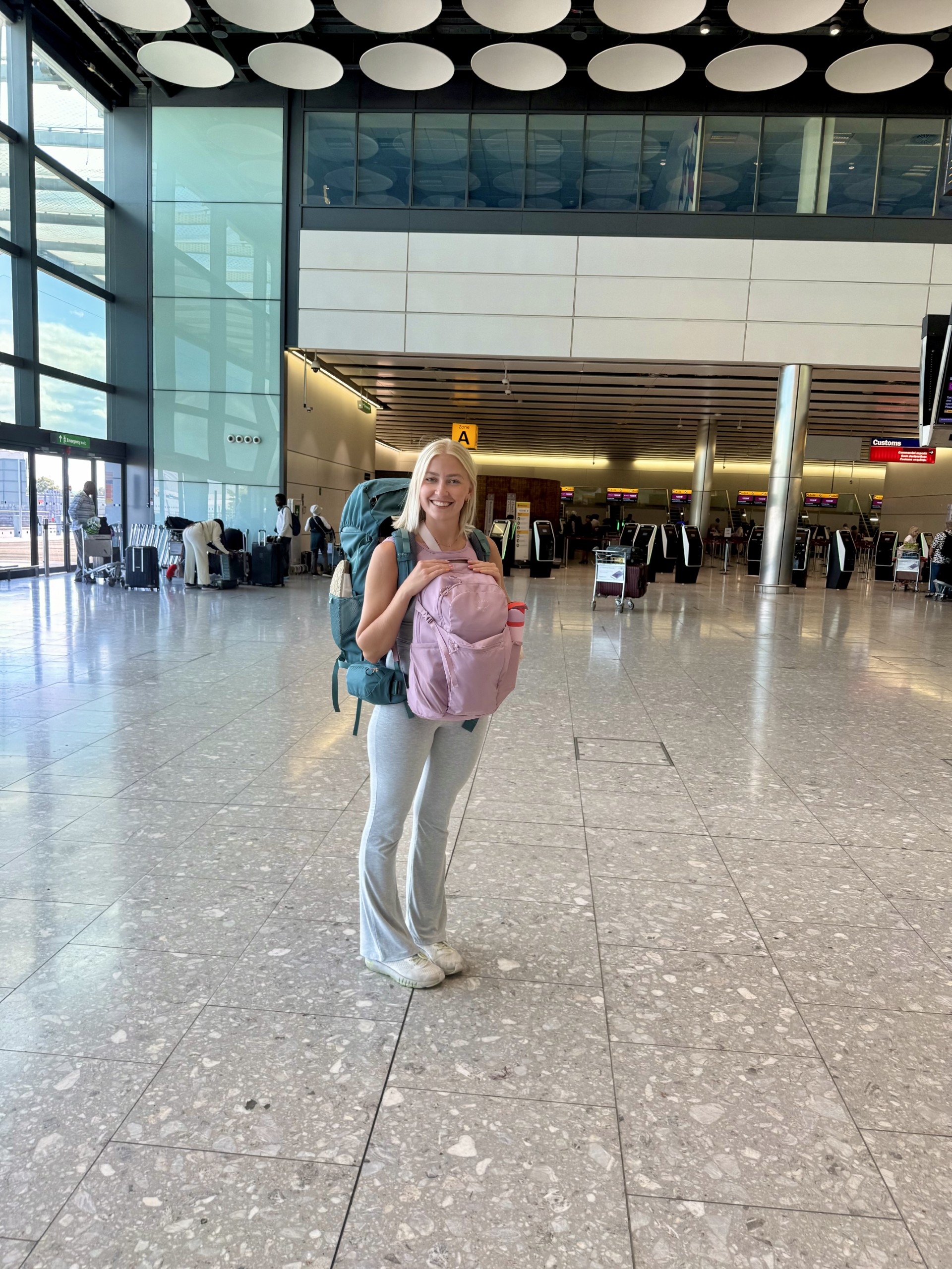 A blonde woman stands in an airport, with a blue backpack on her back and a pink bag on her front.