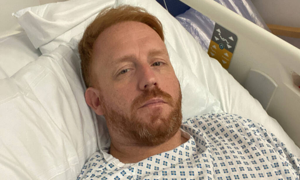 A red-haired man holds a camera and stares tiredly at it after ken butt surgery