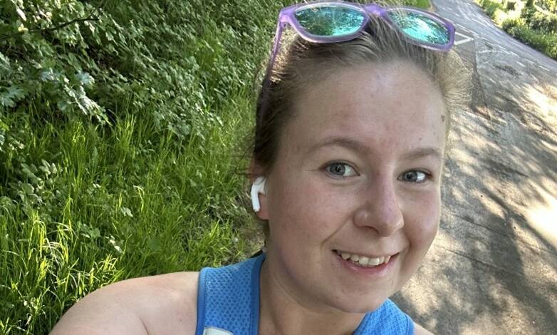 A woman smiles at he camera out on a run, wearing a vest and sunglasses