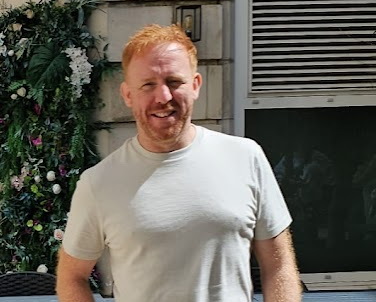 A red-haired man in a white t-shirt stands in front of a table and chairs with his hands in his pockets.