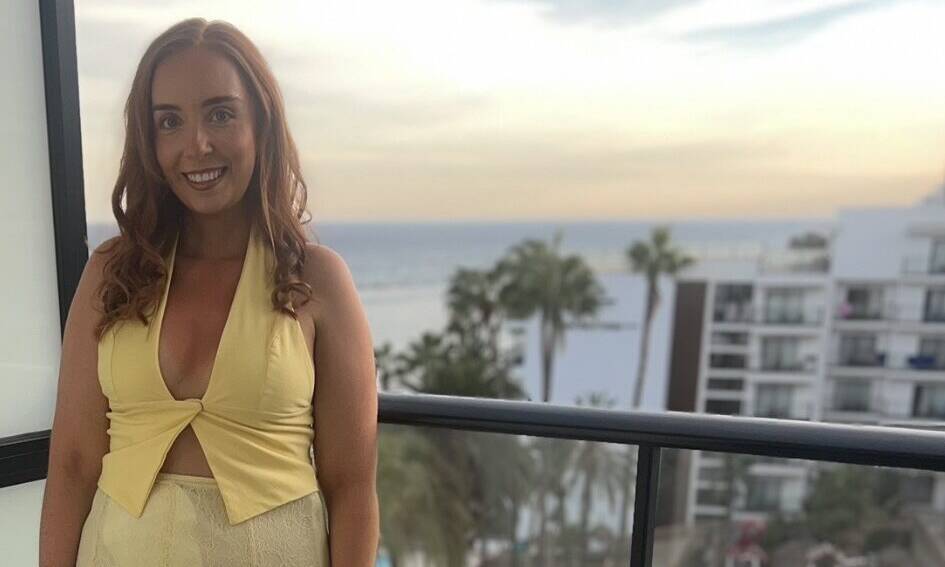 A woman poses in a yellow outfit on a balcony overlooking a pool