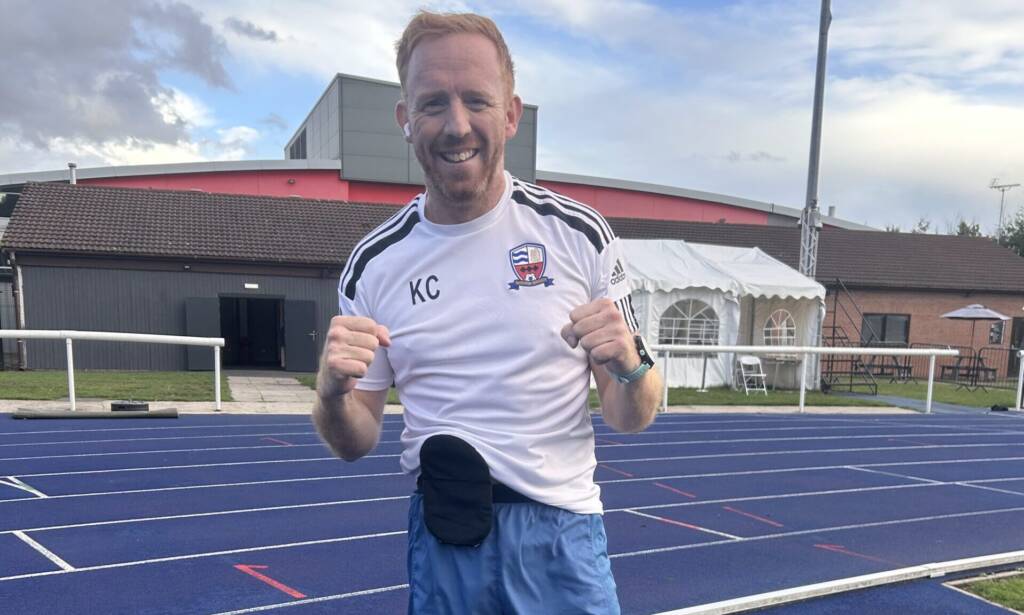 A red-haired man poses with his fists up, dressed in running gear while standing on a blue running track
