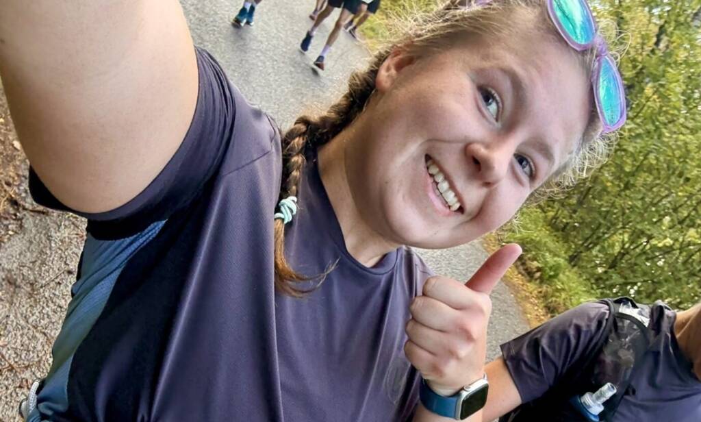A woman standing in a woods, giving a thumbs up while displaying her stoma bag