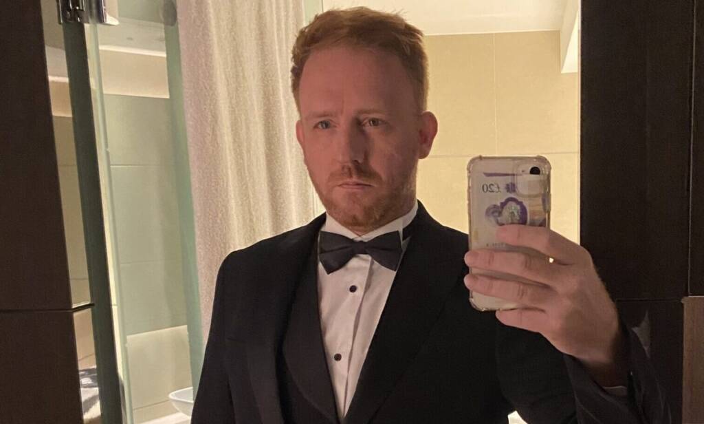 A ginger-haired man wearing a black tuxedo takes a mirror selfie before an event
