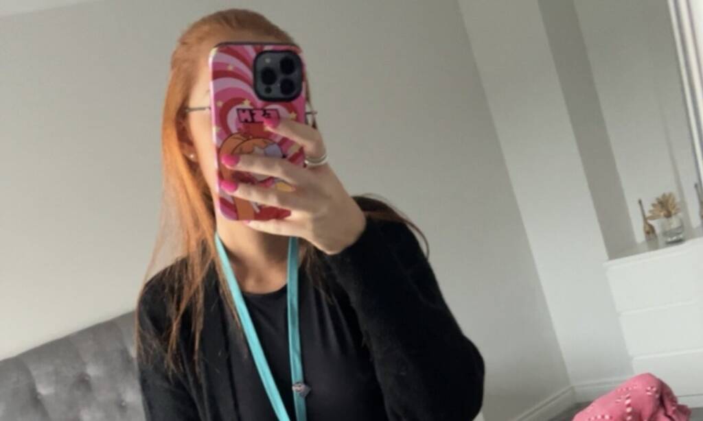 A woman takes a selfie in front of a mirror, standing in front of a bed