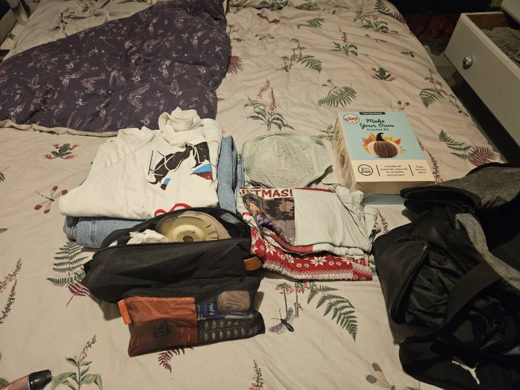 Folded clothes, a crochet kit, toiletries and a bag with stoma products inside sit on a dragonfly bed spread